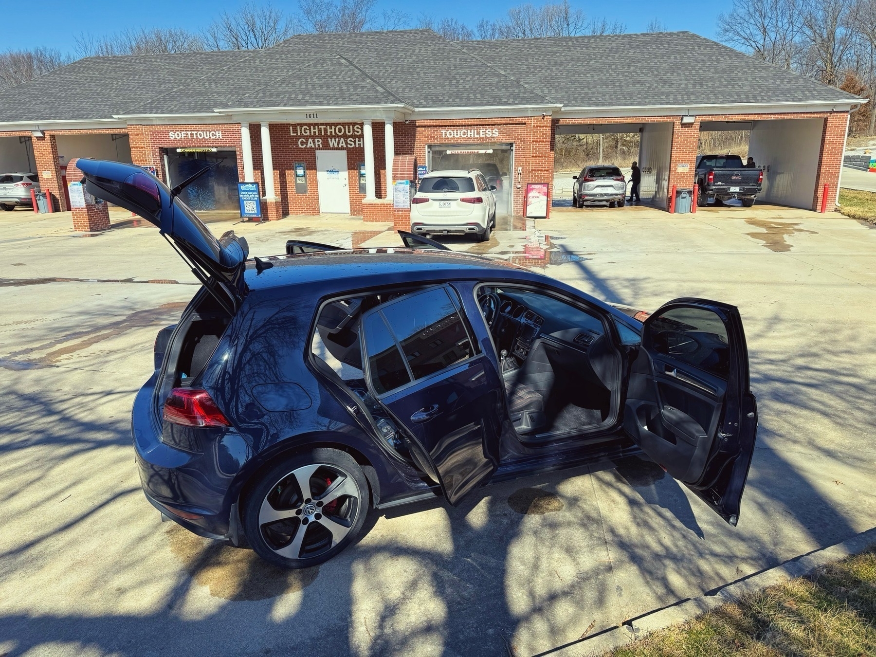 A blue hatchback car with its doors open in the foreground, parked at a car wash facility with "SOFT TOUCH" and "TOUCHLESS" service bays in the background.